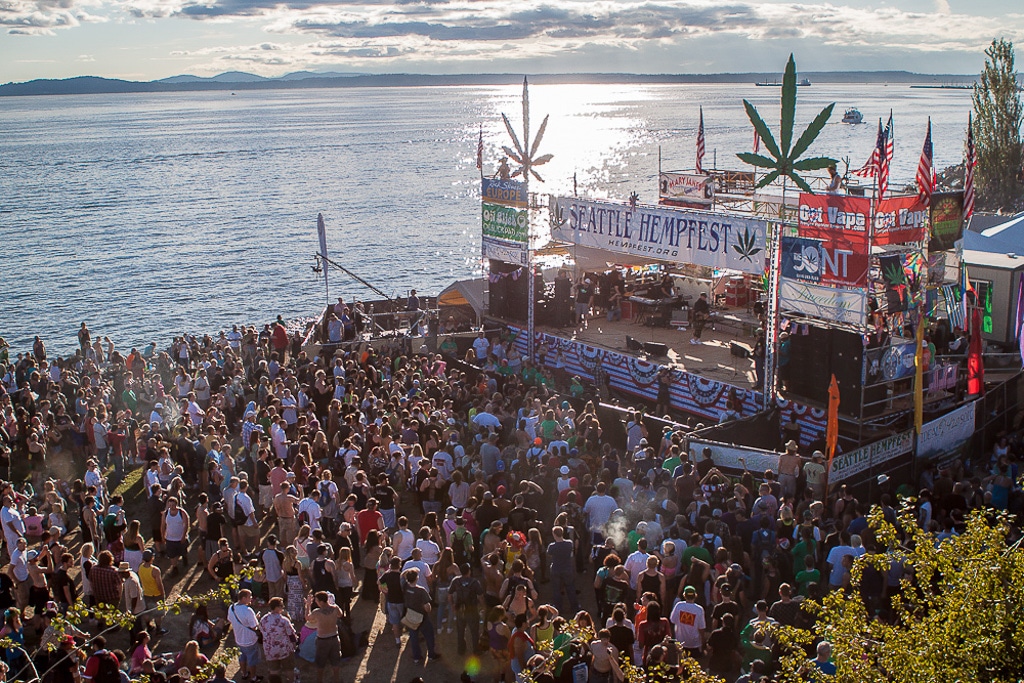 Photo of a large crowd at the annual Seattle Hempfest. There is a large crowd gathered in from of a stage. The sun is setting over Elliot Bay in the background.