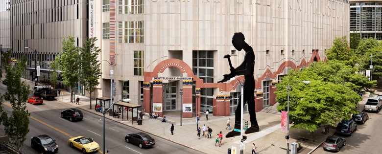 Photo of the Seattle Art Museum exterior. The image shows the front of building from across the street. The iconic Hammer Man sculpture is prevalent.