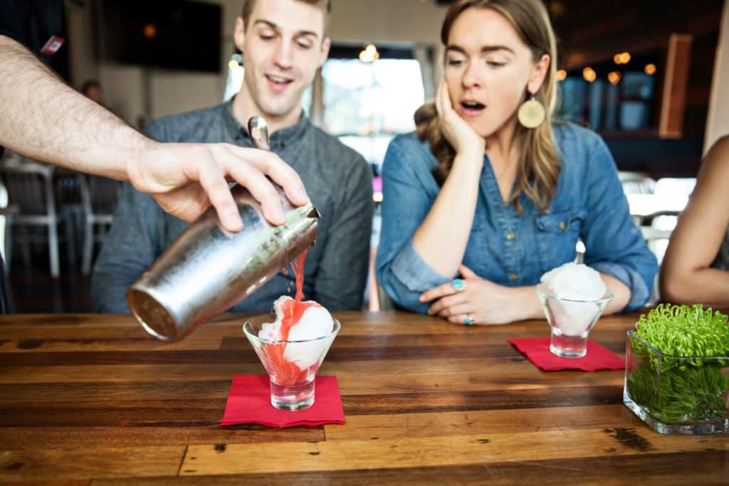 Guests watch with amazement as a cocktail is being poured in front of them. The pink cocktail is being poured over spun sugar as the sugar dissolves.