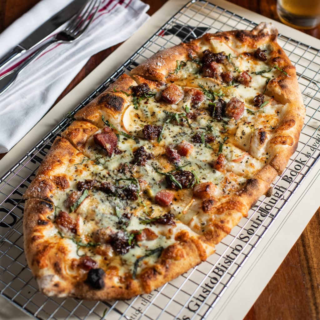 Photograph of a sourdough pizza sitting on a metal trivet atop a wooden table. The pizza has a golden crust and is topped with Applewood smoked slab bacon lardons, Rogue River blue cheese, Gala apple, and fig jam.