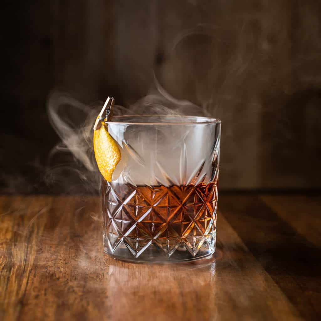Photograph of the Perfect Smoked Manhattan cocktail at Von's 1000 Spirits. The dark brown whiskey cocktail is in a diamond-shaped-etched glass with smoke coming out. There is an orange peel clipped to the outside of the glass for garnish.