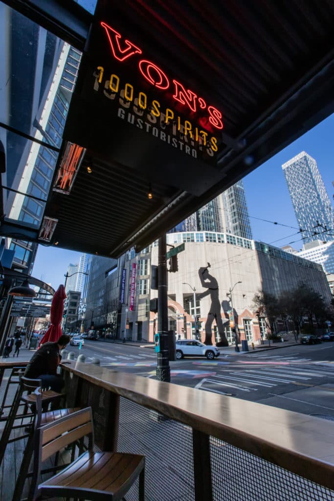 Image of Von's 1000 Spirits street-side front patio, looking out onto the corner of 1st and University in Downtown Seattle. The view is of the Seattle Art Museum and the giant statue of a hammering man on the corner. Other tall buildings can be seen behind the museum.