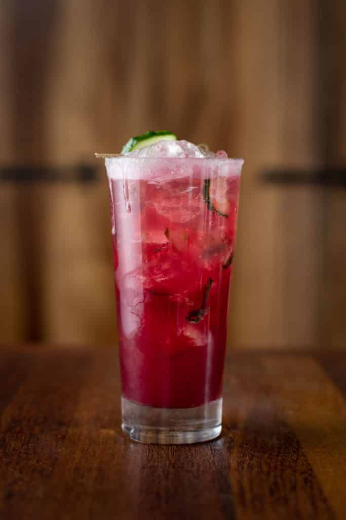 A refreshing iced purple drink sits on a wooden table. It has drips coming down the side of the tall glass and there are slices of cucumber throughout the drink that can be seen through the glass.