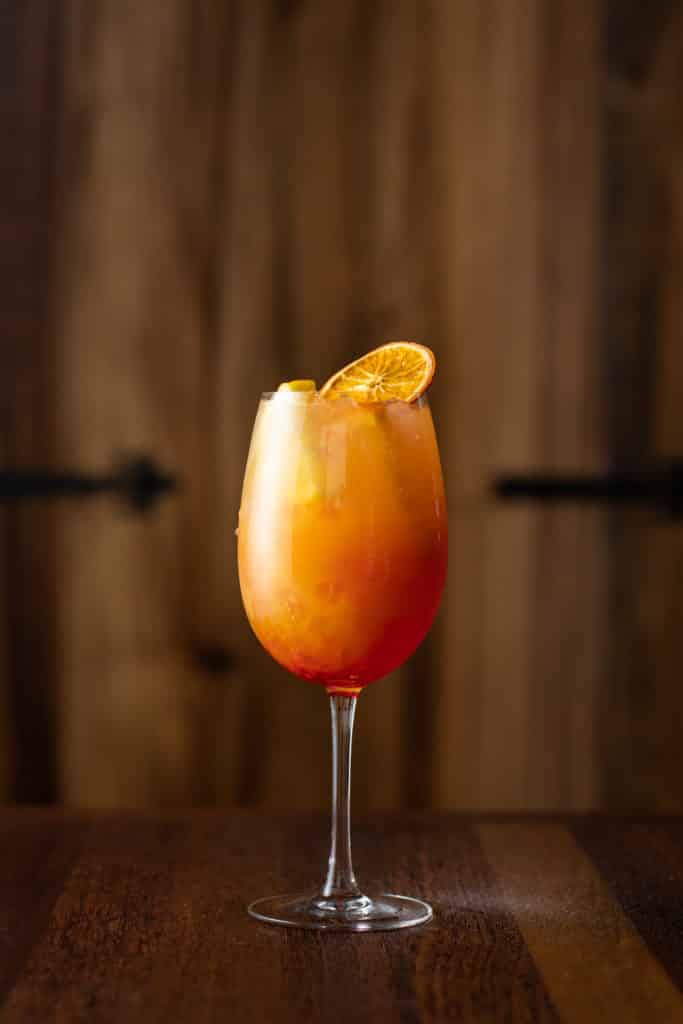 A bright and fruity looking cocktail sits in a wine glass on a wooden table. It is Orange with a red base and has a wheel of dehydrated orange slice on top.