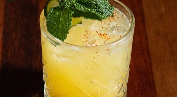 A bright yellow iced cocktail sits on a wooden table in an etched glass. There is a slice of cucumber and a large sprig of mint on top with a sprinkle of red spice.