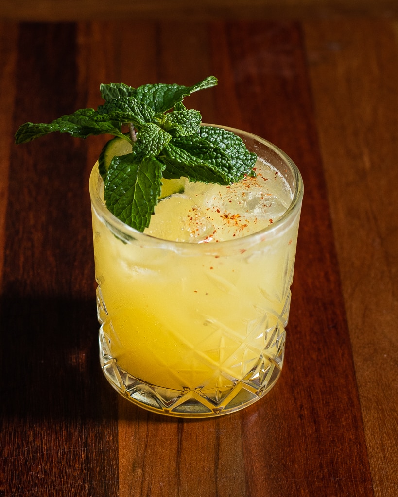 A bright yellow iced cocktail sits on a wooden table in an etched glass. There is a slice of cucumber and a large sprig of mint on top with a sprinkle of red spice.