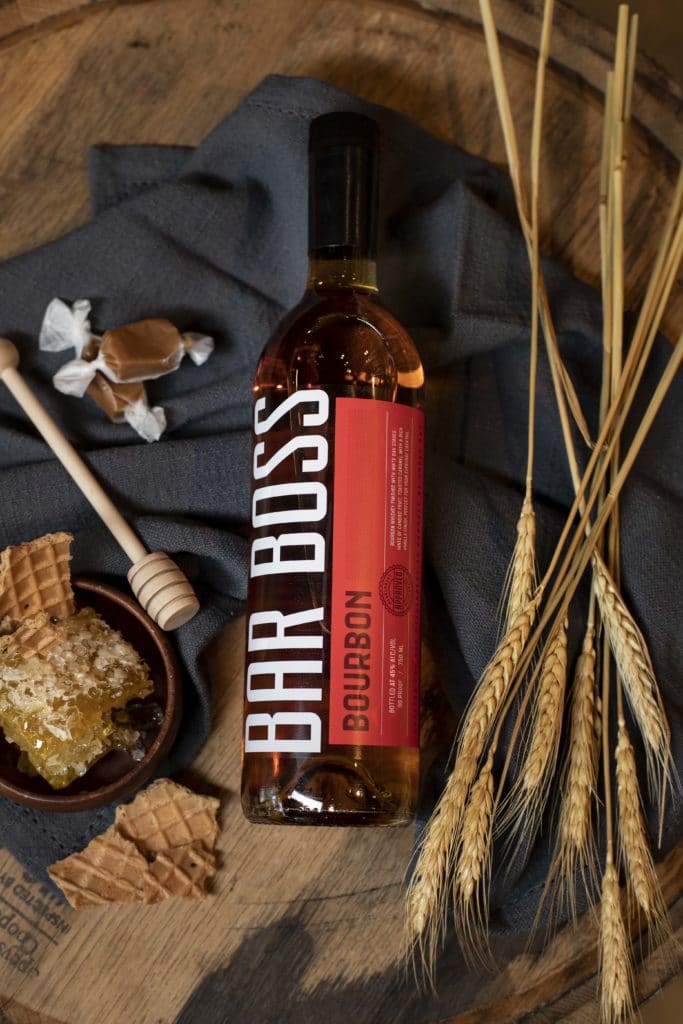 Image of a bottle of Bar Boss Bourbon sitting atop a wooden barrel. The bottle is lying flat on top of a dark blue cloth napkin. The scene around the bottle is decorated with dried wheat, soft caramel candies, honey comb, and waffle cone pieces.