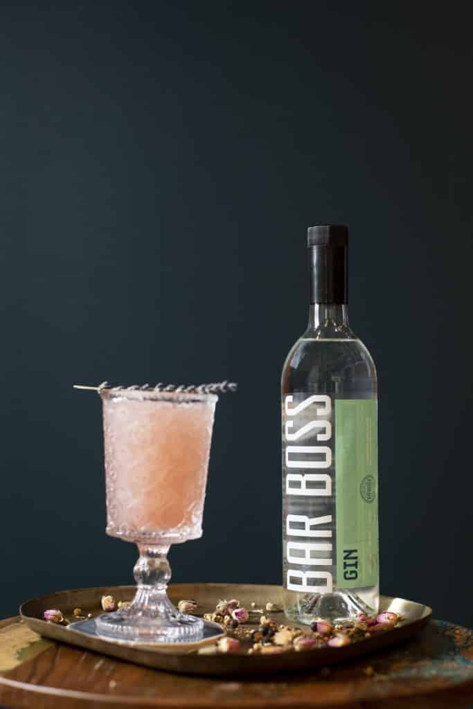 Image of a bottle of Bar Boss Gin sitting on top of a copper tray in front of a dark blue wall. There is a blush pink drink in an etched tall glass next to the bottle. There are dried rose and coriander sitting on the copper tray.  