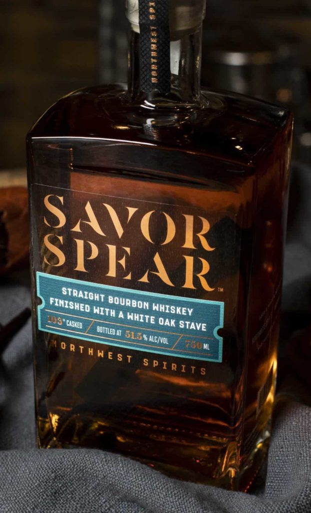 Warm image of a whiskey bottle sitting on a wooden barrel. The Bottle reads “Savor Spear Straight Bourbon Whiskey”. The bottle is sitting atop a blue napkin. The surface around the bottle is adorned with a lit cigar, two soft caramel candies, two vanilla beans, and a leather notebook.