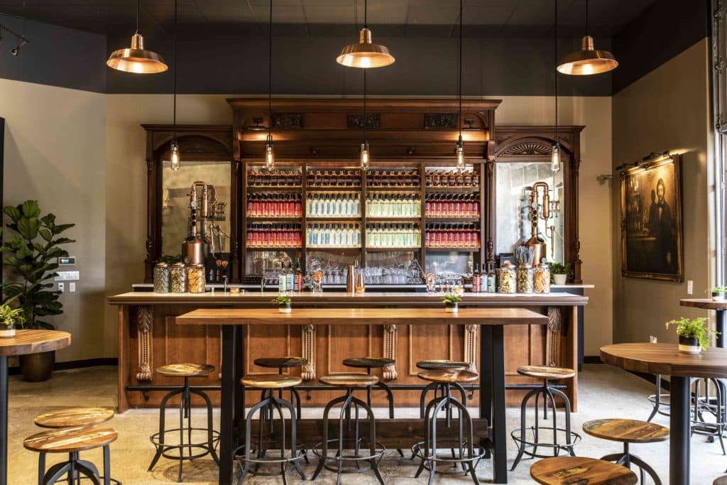 Image of an inviting tasting room. There is an impressive selection of liquor bottles lined up neatly on a Mahogany wooden display behind a white bar top. There are a few wooden tables with wooden stools. There is an industrial, yet warm feeling in the space. 