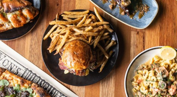 bird's-eye-view image of a table full of delicious food. From top to bottom there Is a salad, meatball sub, chocolate dessert, burger with fries, Mac and cheese, pizza, and clams.