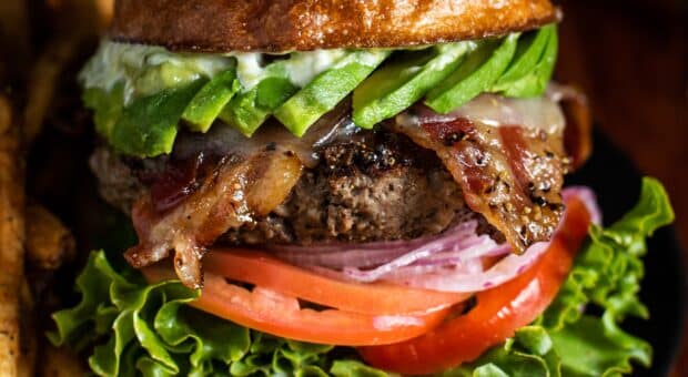 Tantalizing image of a juicy looking burger with a side of golden, crispy fries. The burger has fresh sliced avocado, peppered bacon, juicy tomatoes, red onion, and green leaf lettuce. It sits comfortably in a homemade golden sourdough bun.