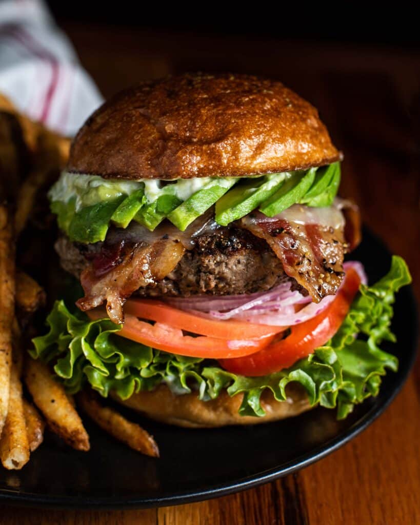 Tantalizing image of a juicy looking burger with a side of golden, crispy fries. The burger has fresh sliced avocado, peppered bacon, juicy tomatoes, red onion, and green leaf lettuce. It sits comfortably in a homemade golden sourdough bun. 