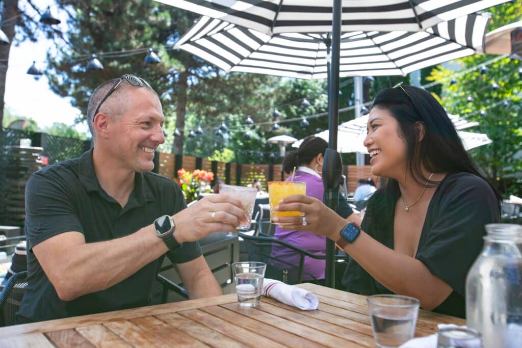 Two people have cocktails in their hands as they "cheers" each other and smile. They are sitting on a nice outdoor patio area that looks inviting.