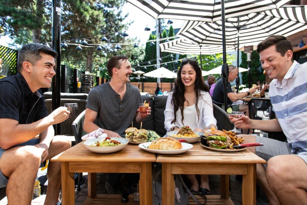 A group of 4 friends sit around an enticing table of food and cocktails as they all smile at one another. They are sitting on a nice outdoor patio area that looks inviting.