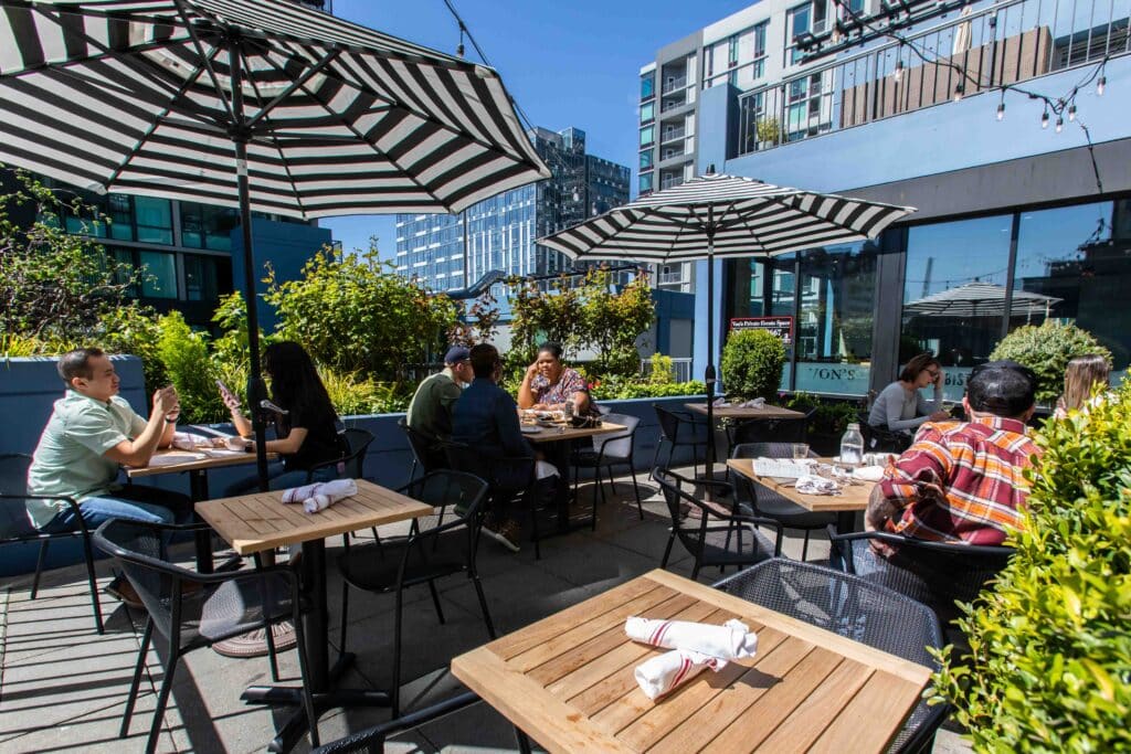 Image of a sunny patio space at Von's 1000Spirits in Downtown Seattle. There are a few tables set with silverware, awaiting guests. There are a few tables with seated guests enjoying themselves. Von's can be seen in the background as well as some of the Seattle skyline.