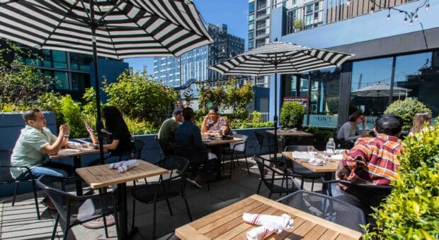 Image of a sunny patio space at Von's 1000Spirits in Downtown Seattle. There are a few tables set with silverware, awaiting guests. There are a few tables with seated guests enjoying themselves. Von's can be seen in the background as well as some of the Seattle skyline.