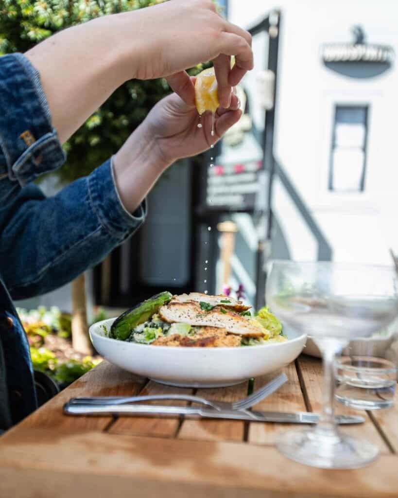 Image of a woman's hands squeezing a lemon onto a Chicken Cesar Salad. The salad sits on a wooden table on an outdoor patio.