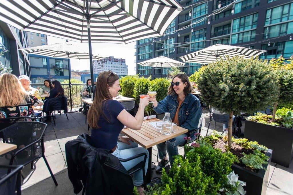 Image of 2 women holding their cocktails up for a "Cheers". They are smiling and having a good time. They are seated on a lovely sunny outdoor patio at Von's 1000Spirits in Downtown Seattle. 