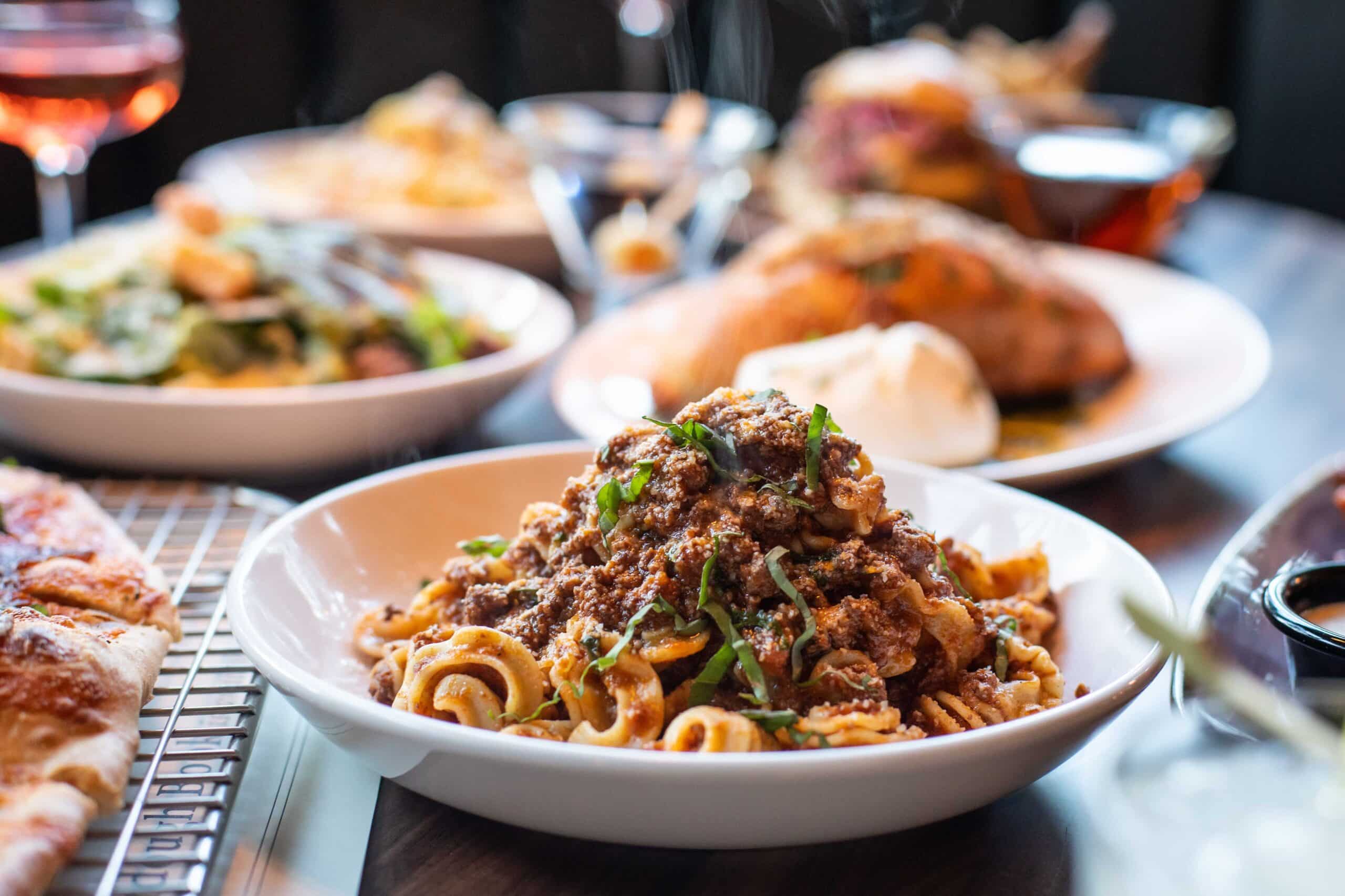 A scrumptious looking plate of pasta bolognese sits on a table among a feast. Steam rises from the dish, evoking a sensory response.