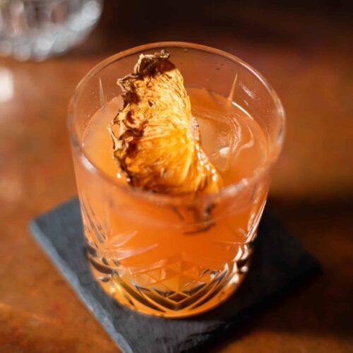 Pineapple Old Fashioned cocktail served in a cut-crystal glass over a large ice cube, garnished with a dried pineapple slice, on a dark slate coaster.